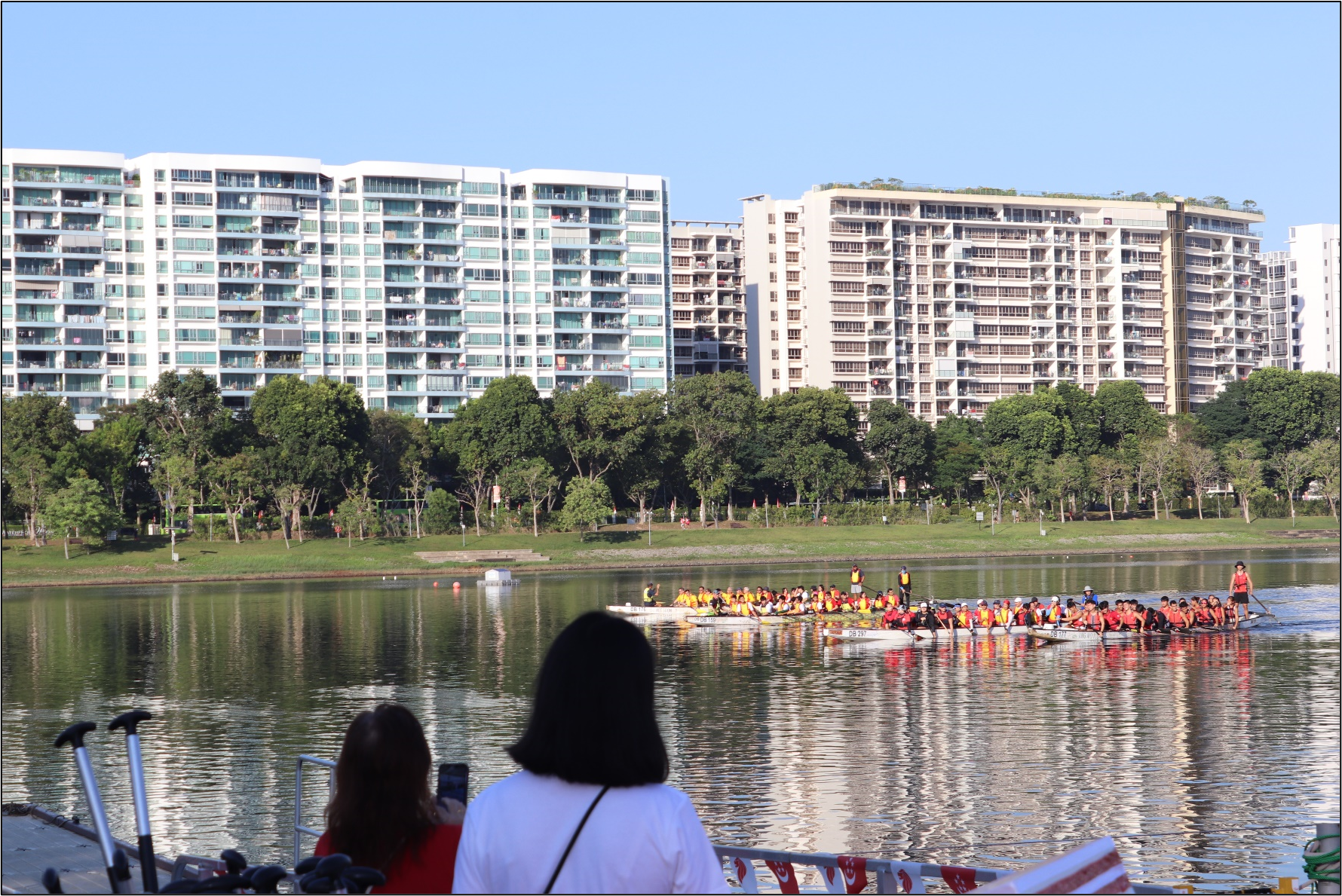 Two spectators watch a dragon boat race on a lake with high-rise apartments and trees in the background.